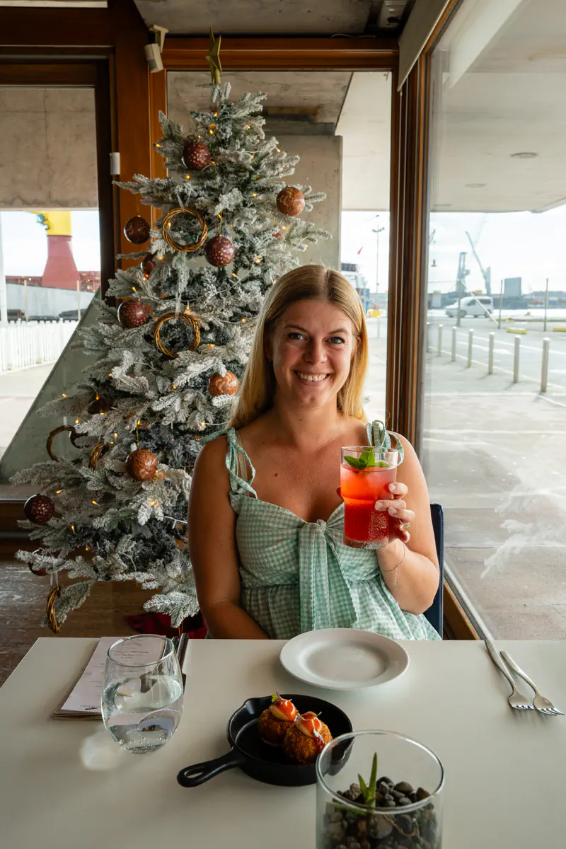 A woman sits at a restaurant table holding a pink lemonade and smiling toward the camera. A decorated Christmas tree and large windows are visible behind her, with small plates of food on the table.