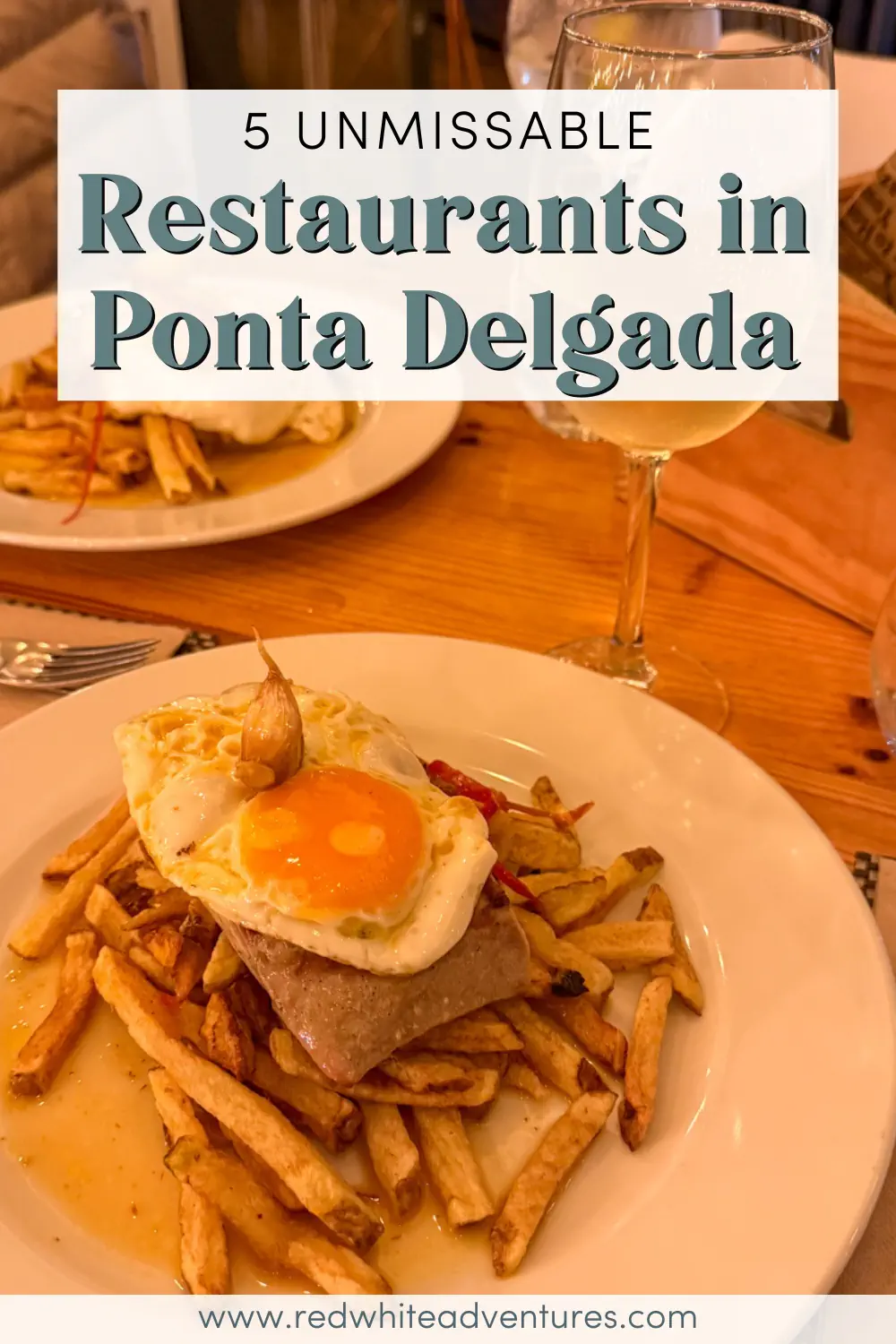 A plate of fries topped with a tuna steak and a sunny-side-up egg sits on a wooden restaurant table beside a glass of white wine. Text over the image reads “5 Unmissable Restaurants in Ponta Delgada.”