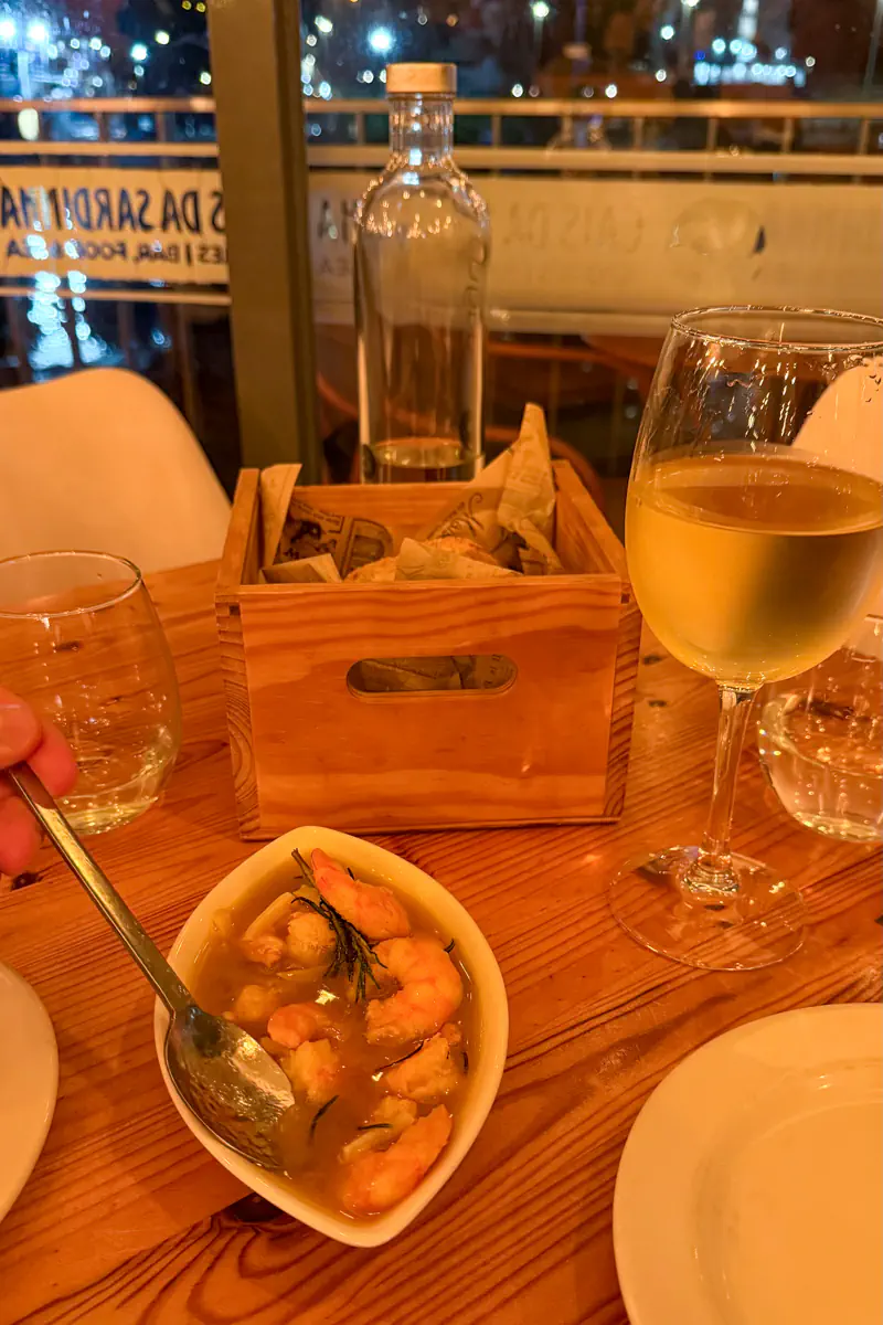 A small bowl of shrimp in sauce sits on a wooden table beside glasses of white wine and water. A wooden box with folded paper and a glass bottle is placed near the window in the background.