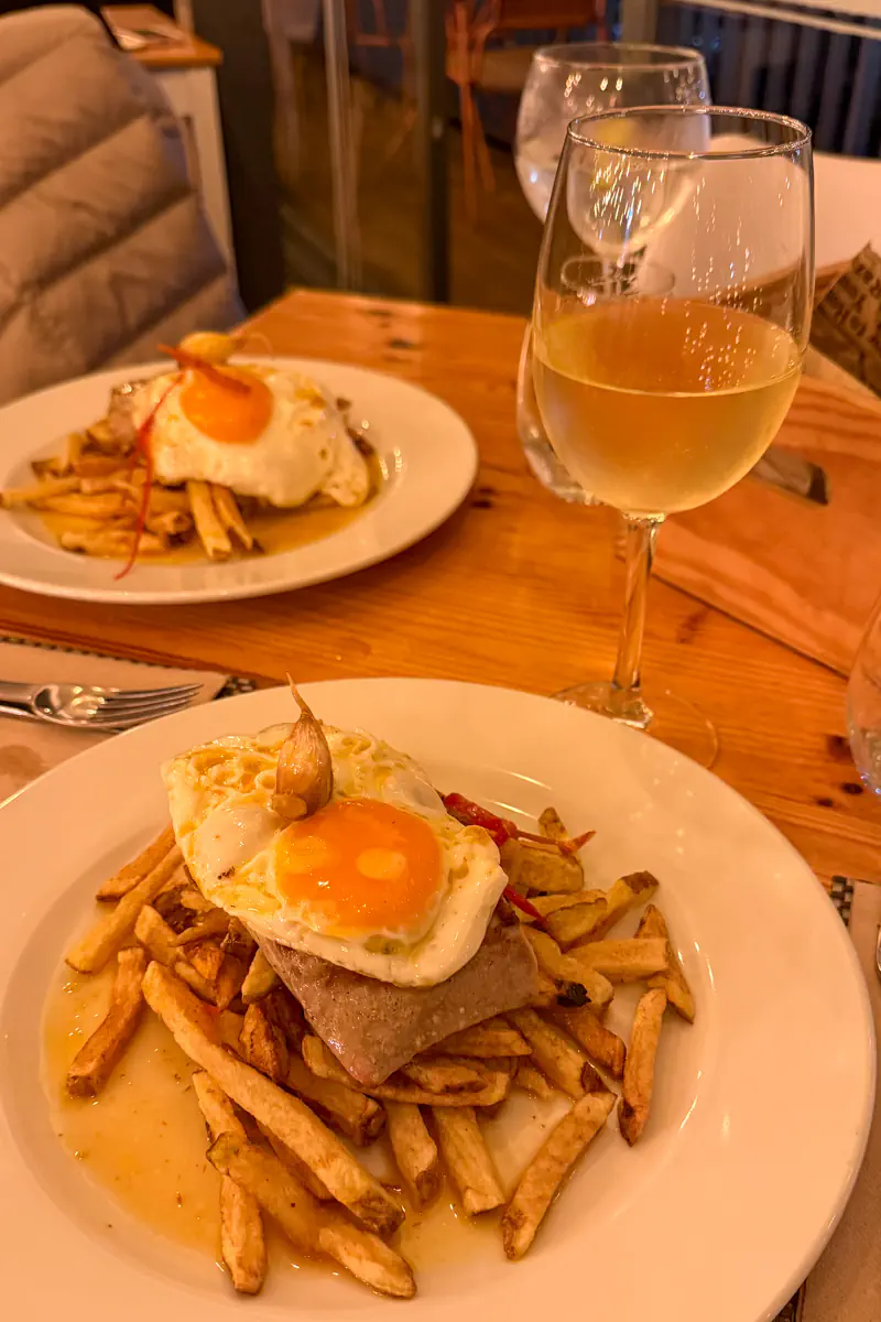 A tuna steak topped with a fried egg is served over fries on a white plate, with a glass of white wine beside it. Another similar dish is visible across the table in a warmly lit restaurant setting.