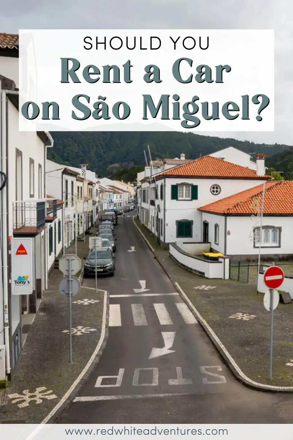 A narrow street lined with white houses and red tile roofs runs through a town on São Miguel Island, with parked cars along the road. Text overlay reads, “Should You Rent a Car on São Miguel?”