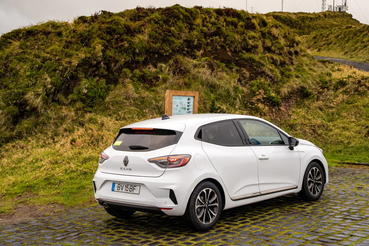A white rental car is parked on a cobblestone pull-off beside a grassy hillside on São Miguel Island. A small roadside signboard and sloping green terrain surround the vehicle.