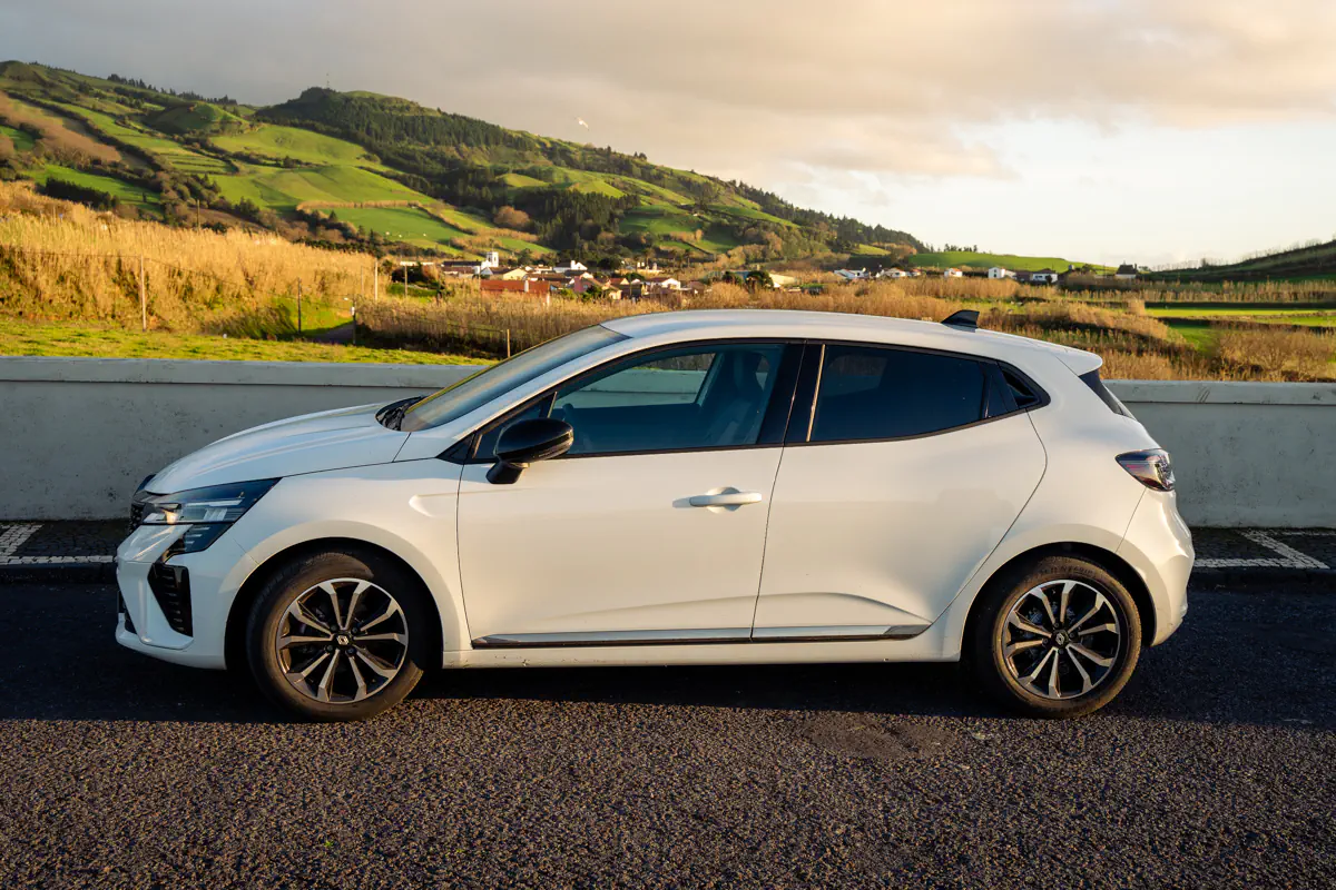 A white rental car is parked roadside with rolling green hills and small villages in the background. Warm sunlight highlights the Azores countryside.