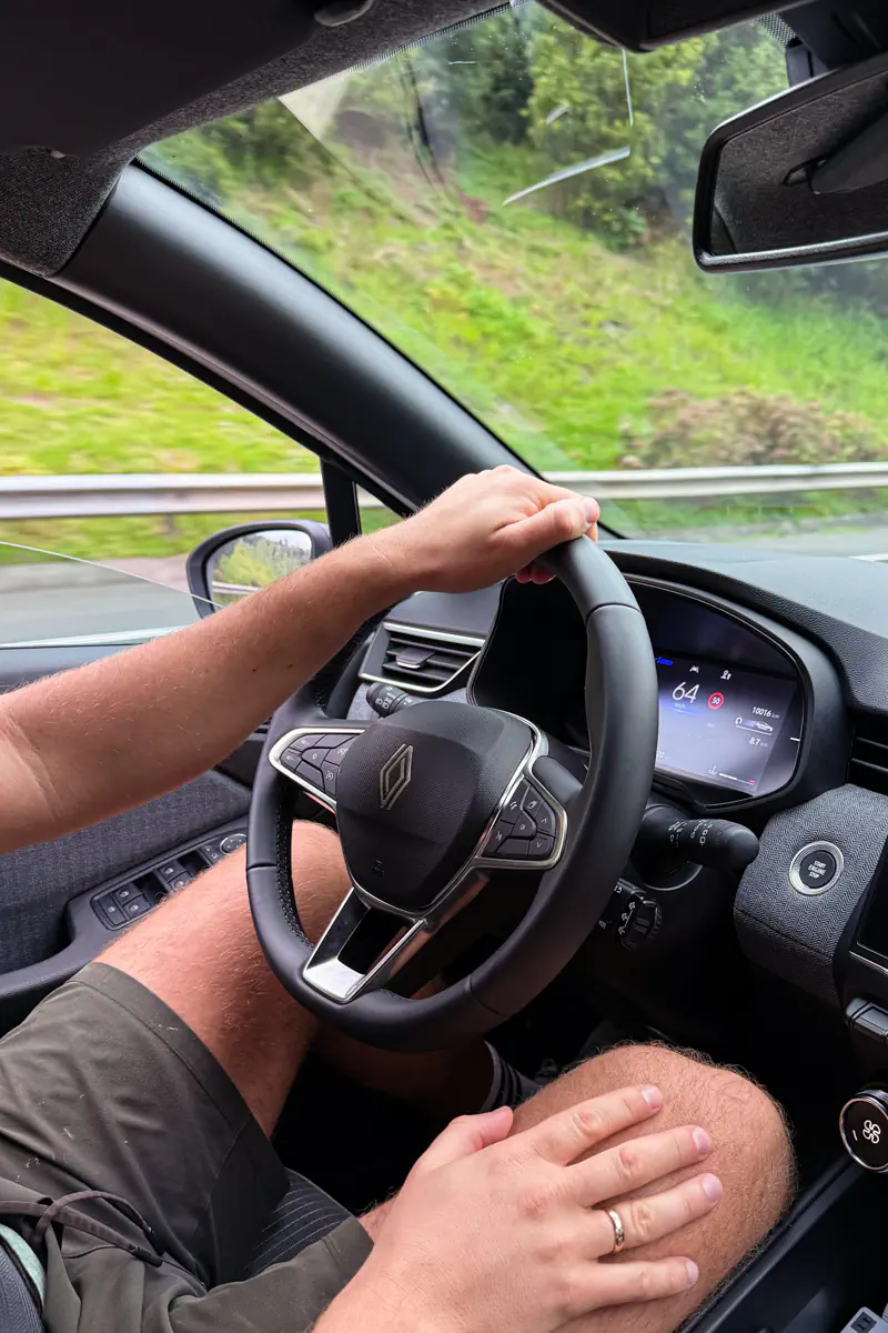 A driver’s hands rest on the steering wheel while driving along a scenic road on São Miguel Island. Green hills and guardrails are visible through the windshield and window.