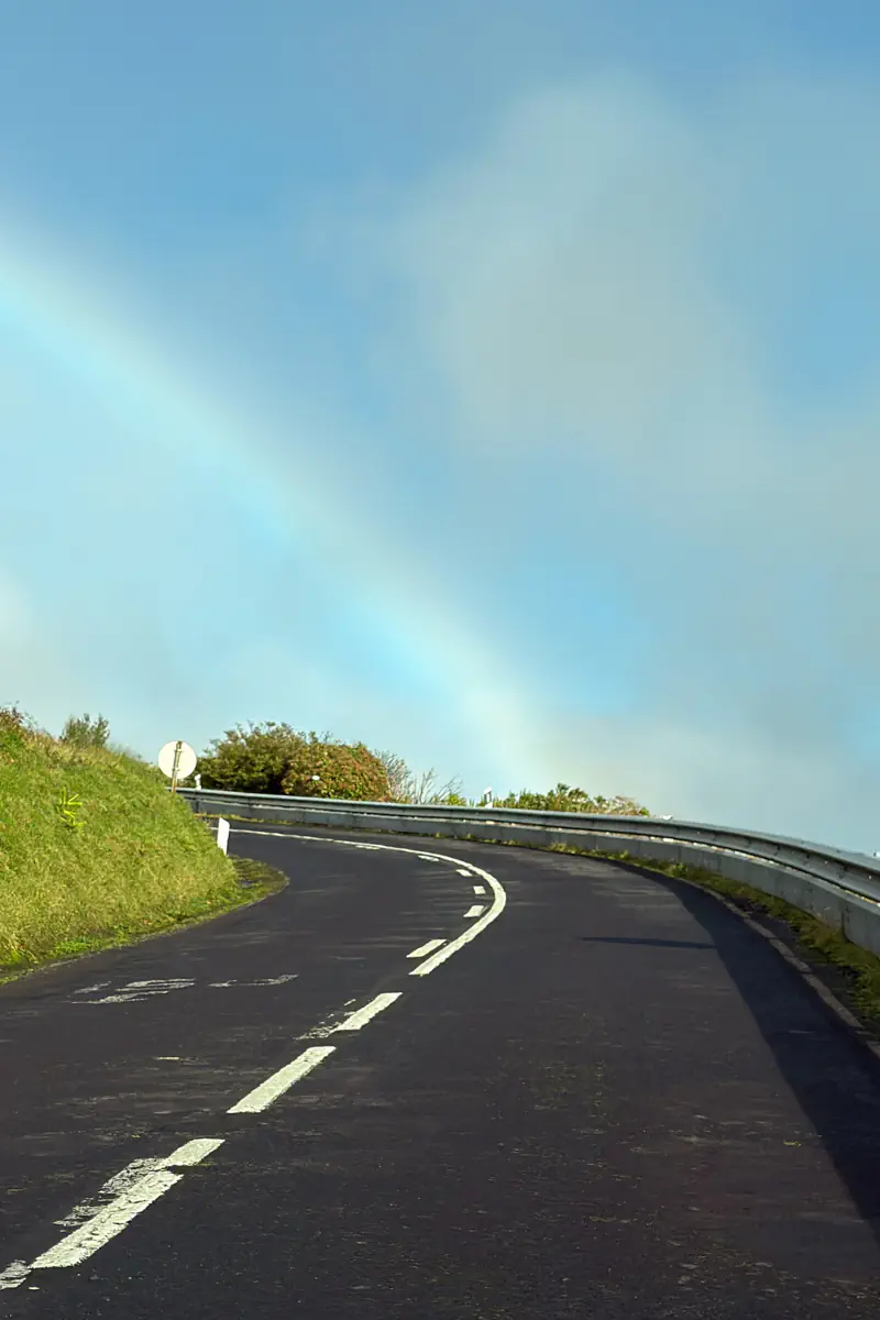 A curved mountain road on São Miguel Island sits beneath a soft blue sky with a faint rainbow visible above the bend. Green hills and guardrails frame the scenic drive.