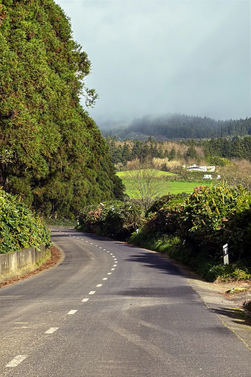 A quiet country road curves through lush greenery with misty clouds hanging over the hills ahead. The scene captures São Miguel’s moody weather and rural charm.