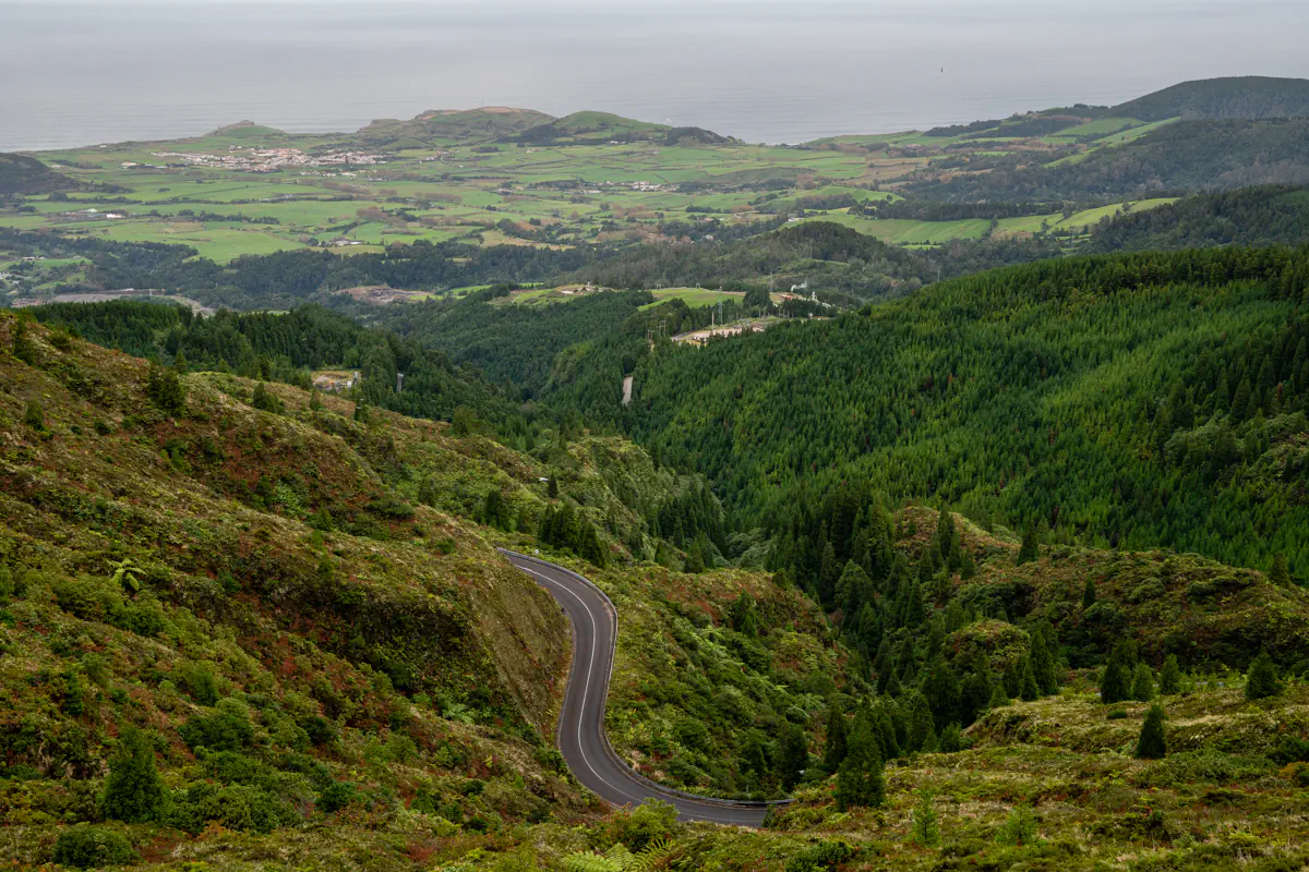 A winding mountain road cuts through lush green hills with farmland and the coastline visible in the distance. The elevated viewpoint highlights São Miguel’s dramatic landscape.