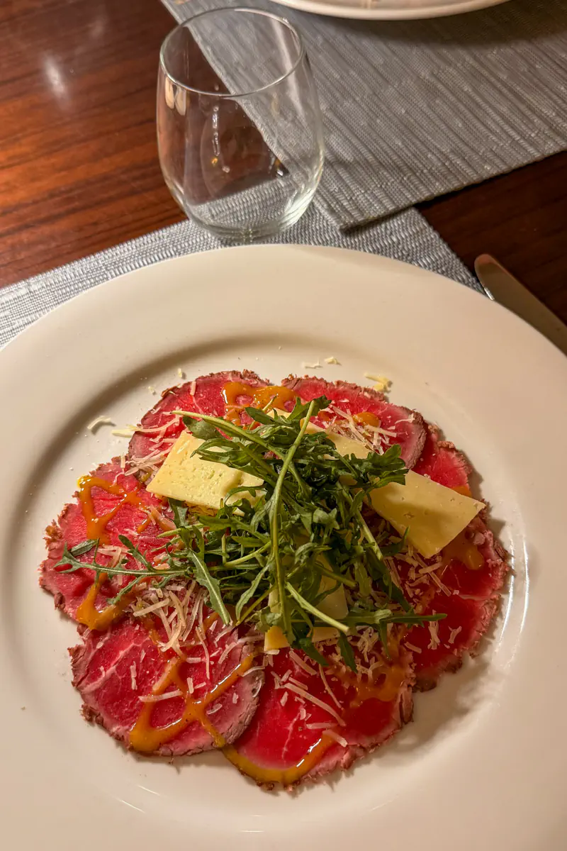 A plate of thinly sliced beef carpaccio is topped with arugula, shaved cheese, and a light drizzle of sauce. The dish is served on a white plate at a restaurant table with glassware nearby.