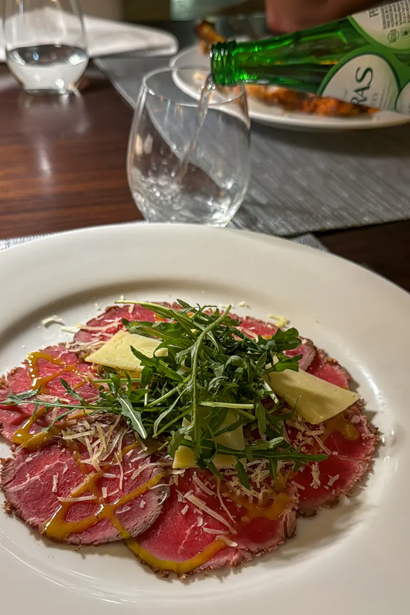 Beef carpaccio topped with arugula and shaved cheese sits on a white plate as water is poured into a glass beside it. The scene is set on a restaurant table with place settings visible.