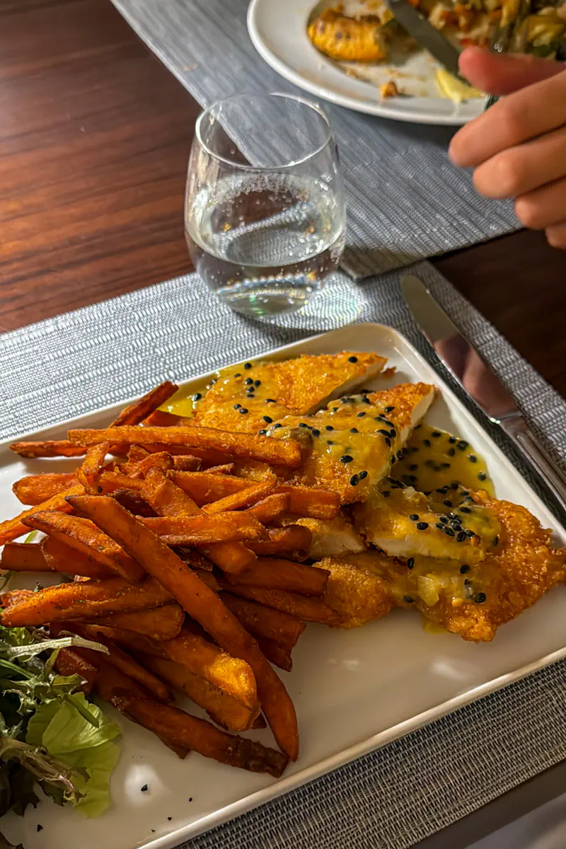 A breaded chicken fillet is served with sweet potato fries and passionfruit sauce on a rectangular white plate. A glass of water and a diner’s hand holding cutlery are visible at the table.