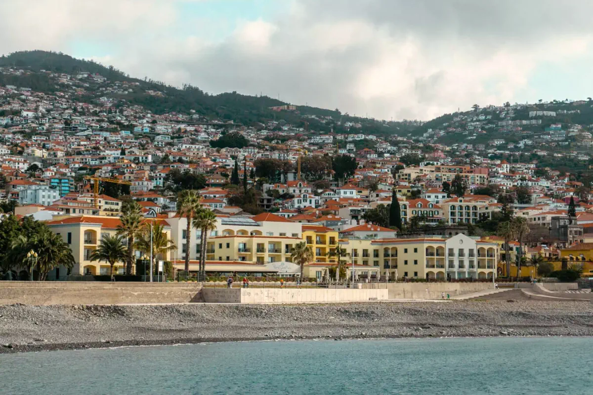 A coastal view of Funchal, Madeira shows colourful buildings with red-tiled roofs stretching up the hillside behind a waterfront promenade. The calm ocean sits in the foreground, with palm trees and low-rise hotels and apartments lining the shore under cloudy skies.