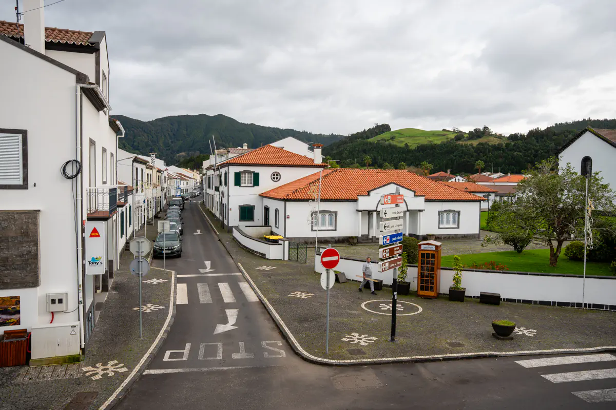A quiet street in a small São Miguel town lined with white buildings and red-tiled roofs. Road markings and directional signs are visible under an overcast sky.