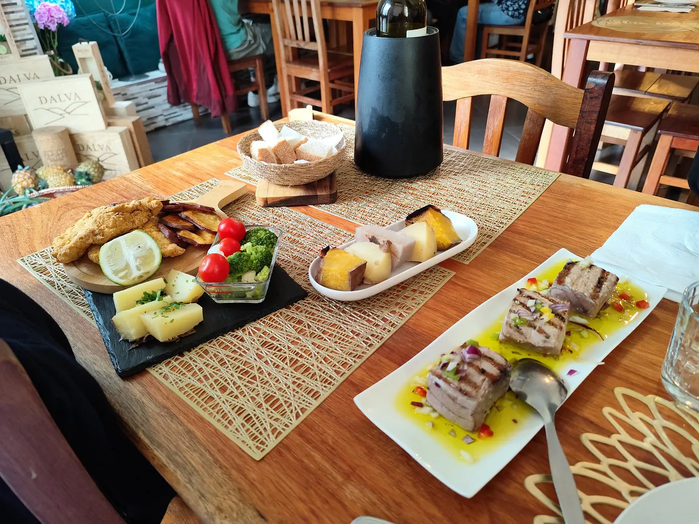 A wooden restaurant table is set with several small plates of food, including grilled fish with olive oil and chopped vegetables, sliced cheese, fried fish, potatoes, and vegetables. A wine bottle in a black cooler, bread basket, and place settings are arranged on the table.