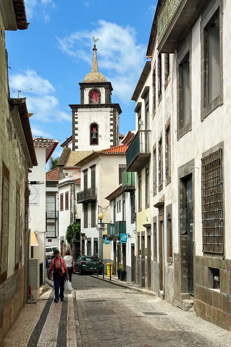 A narrow Portuguese-style cobblestone street lined with traditional buildings leads toward a church tower with a bell and cross at the top. A pedestrian walks along the sidewalk beneath blue skies, with balconies and shuttered windows framing the historic street scene.
