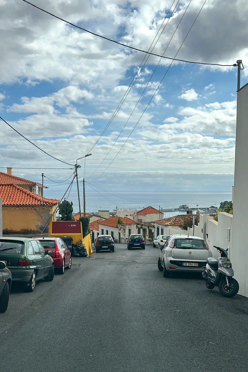 A sloped residential street with parked cars descends toward the ocean in the distance. Red-tiled roofs and low buildings line the road under a partly cloudy sky overlooking the water.