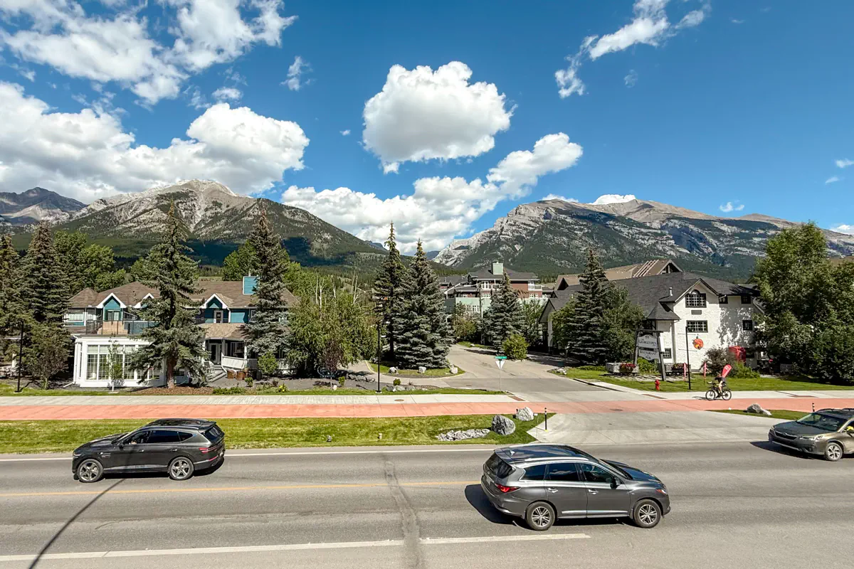 A wide view of Canmore, Alberta, with houses, trees, and a road in the foreground and mountains under a blue sky with scattered clouds.