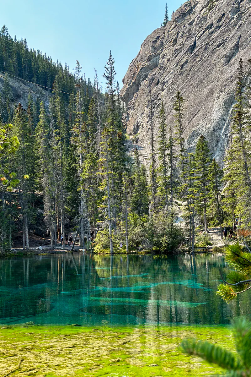 A clear turquoise pond surrounded by pine trees and a rocky cliff, with reflections visible in the water and a few people standing along the shoreline.