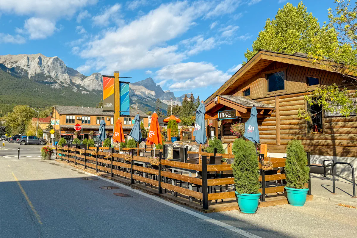A street in Canmore with a wooden patio restaurant, with mountains in the background under a blue but partly cloudy sky.