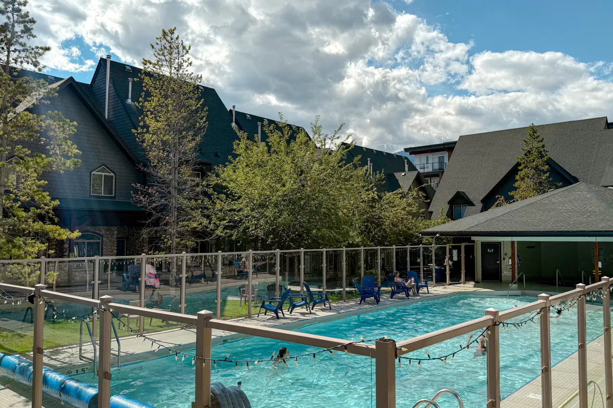 Outdoor pool area at a mountain lodge in Canmore, with lounge chairs, surrounding buildings, and forested scenery under a partly cloudy sky.