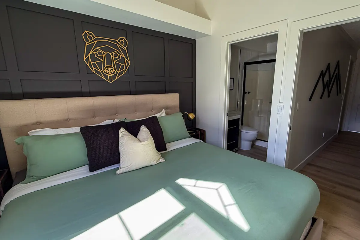 A modern hotel room with a large bed, green bedding, and a geometric bear wall design above the headboard, with an ensuite bathroom visible.