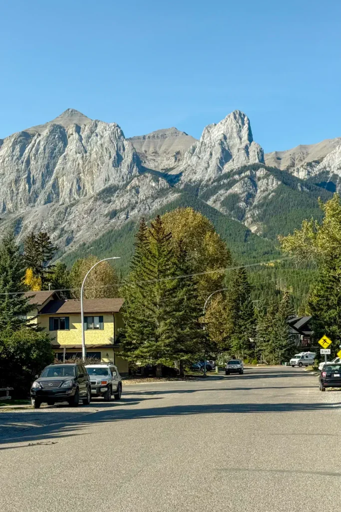 A quiet residential street in Canmore with houses, parked cars, and dramatic rocky mountain peaks in the background.