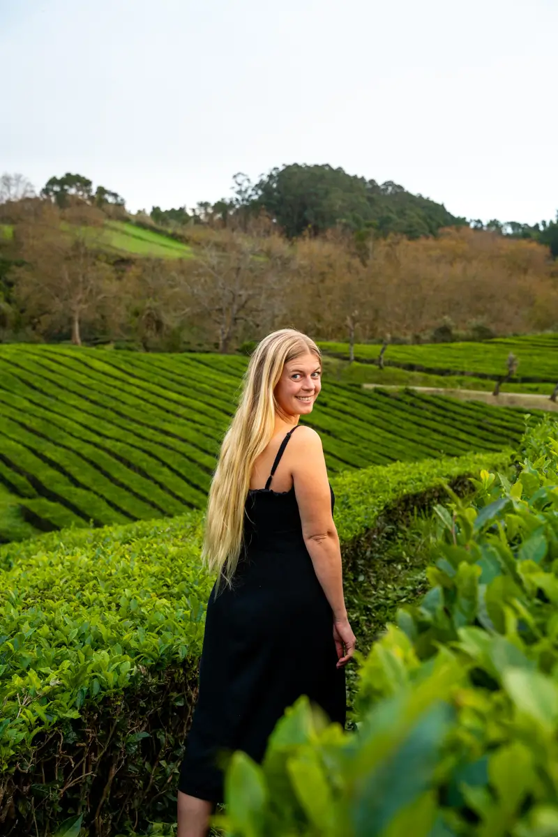 A woman in a black dress standing in tea fields and turning back toward the camera, with rolling green hills behind her.