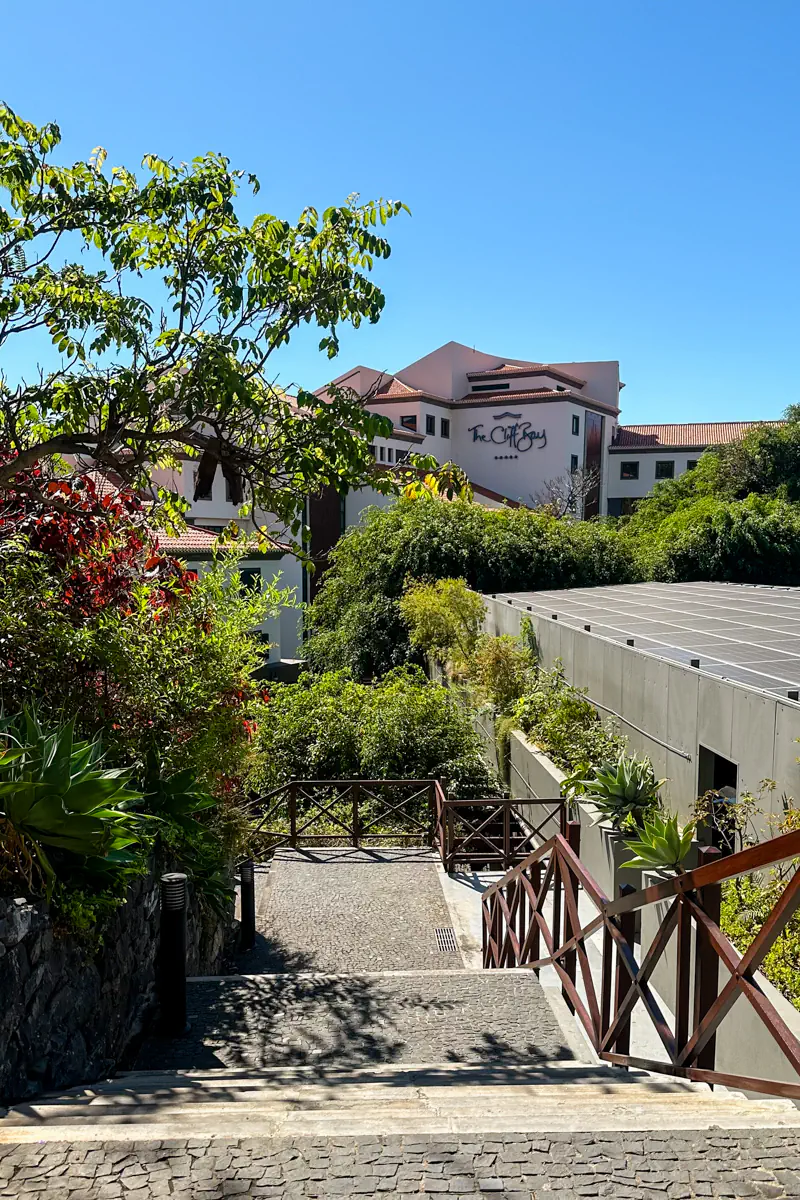 Stone steps with wooden railings lead down through lush greenery toward a large hotel building under a clear blue sky. The hotel sign is visible on the upper facade above the surrounding trees.