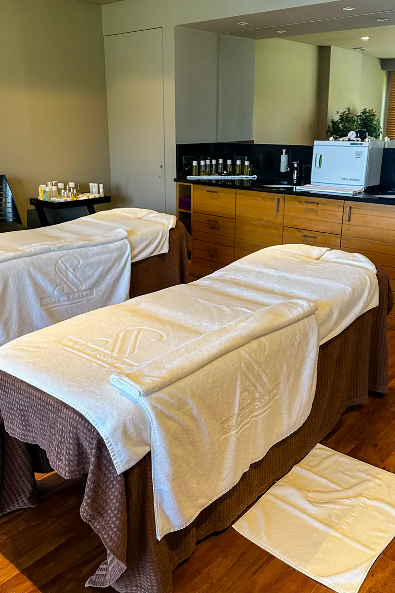 A spa treatment room features two massage tables covered with white towels and folded blankets. Wooden cabinets, skincare products, and soft lighting create a calm, relaxing atmosphere.