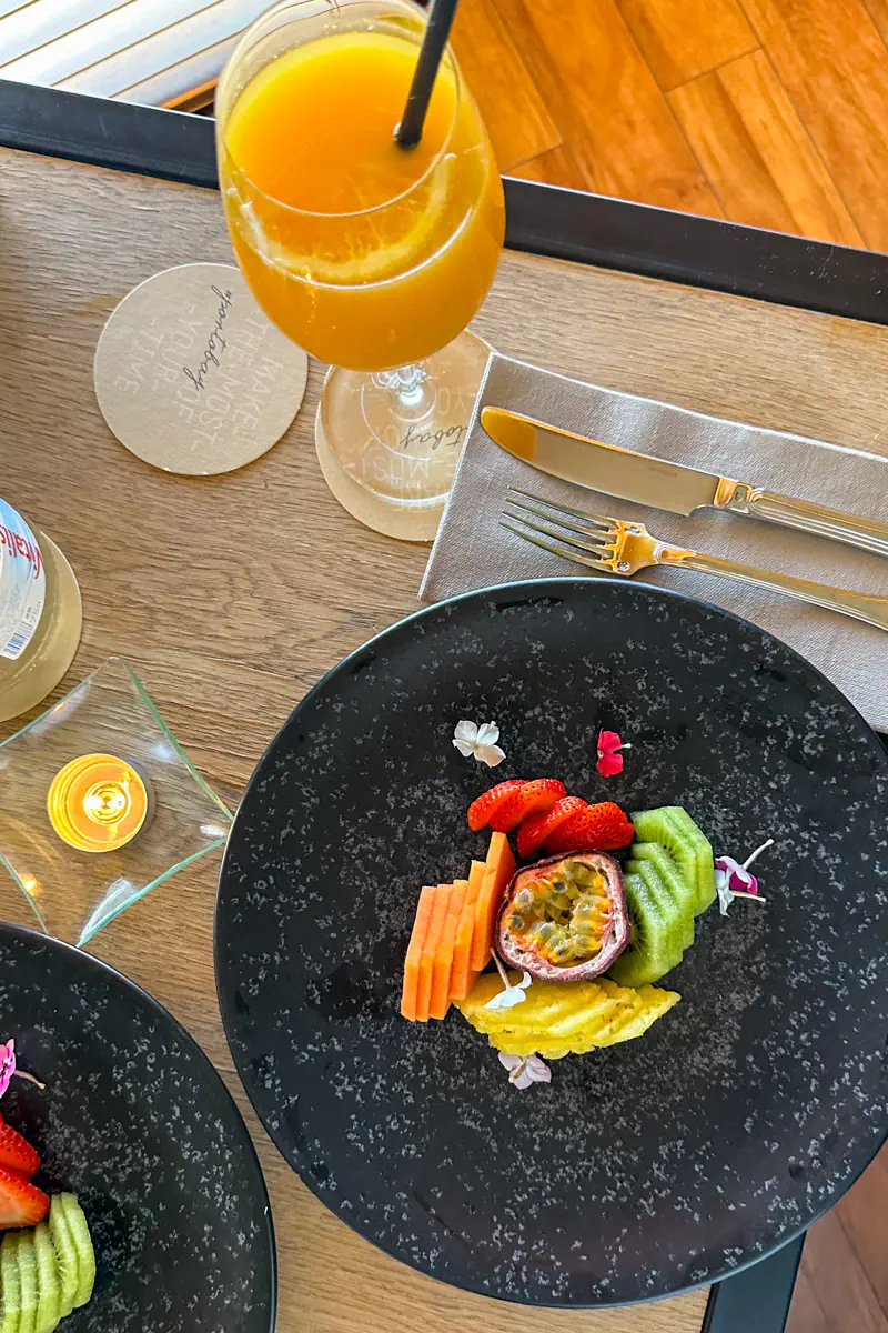 A plate of sliced fresh fruit including kiwi, strawberries, pineapple, and passion fruit is arranged on a dark plate beside a glass of orange juice. A candle and table setting sit on a wooden table in a bright dining area.