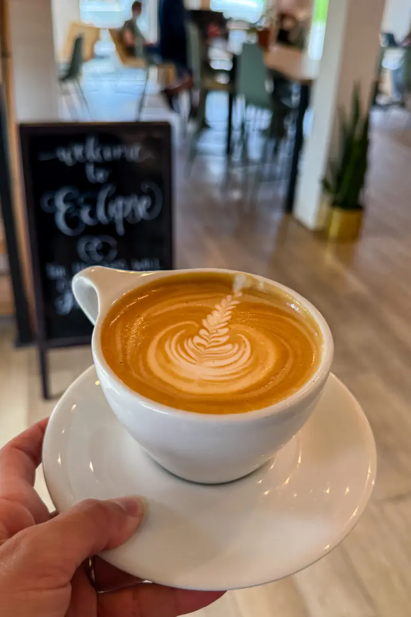 A flat white with latte art held in front of a blurred café interior, with a sign in the background that reads “Welcome to Eclipse.”