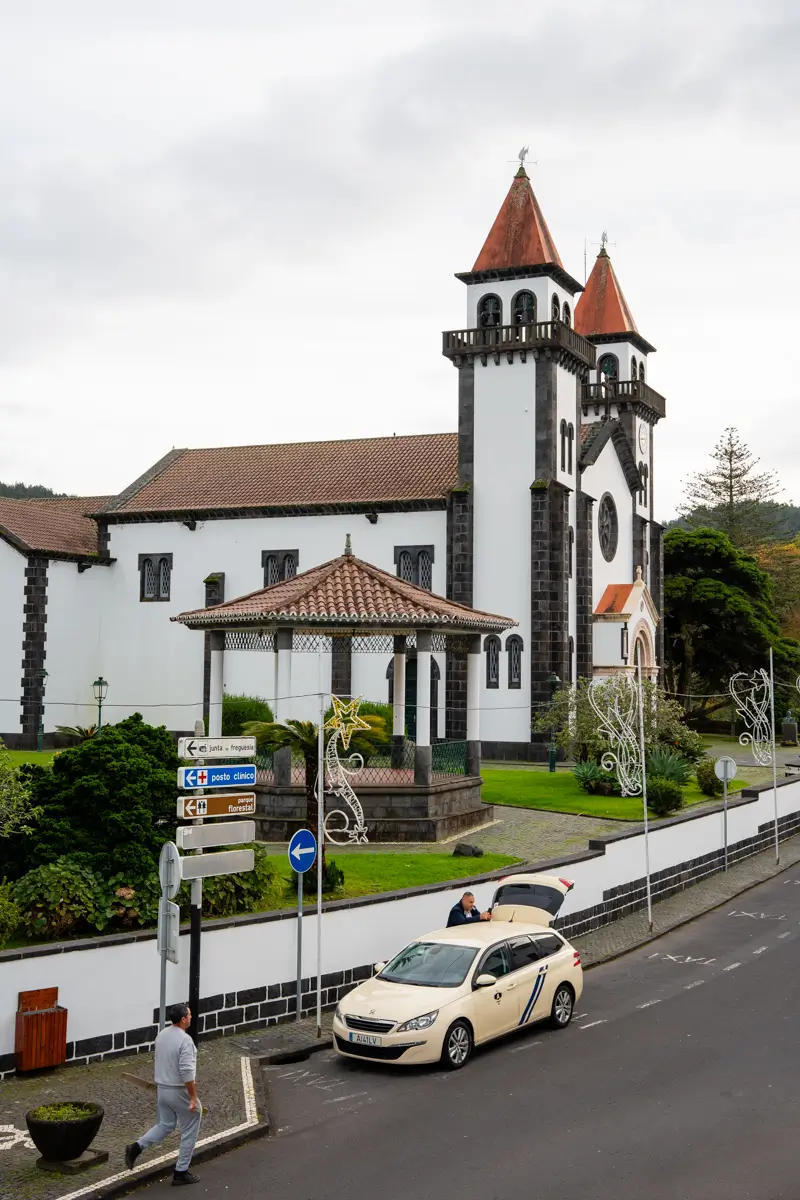 White church with twin towers and red roofs in a small town in the Azores, with a car parked on the street and mountains in the background.