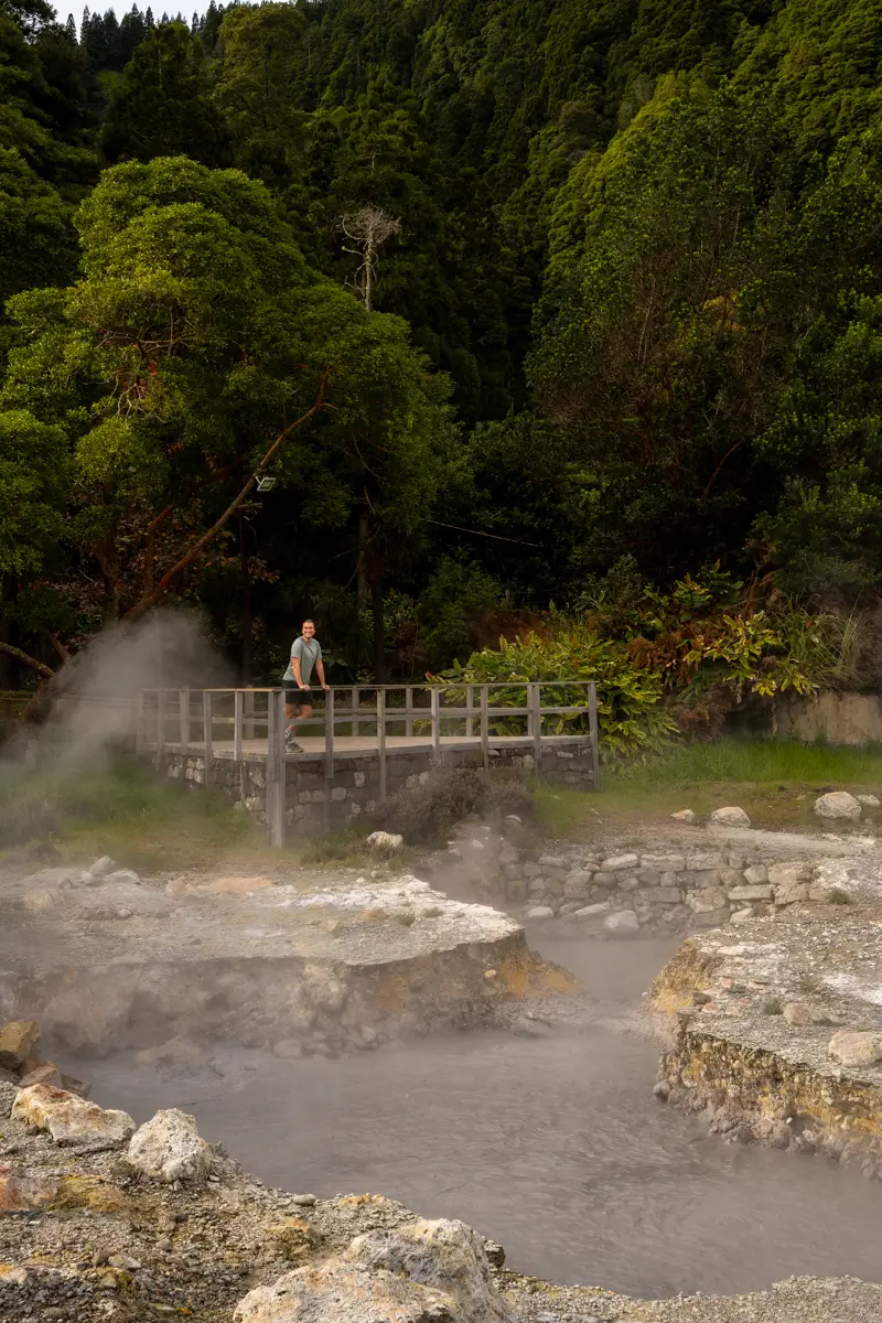 A person standing on a wooden platform above steaming geothermal pools, surrounded by dense green forest in the Azores.