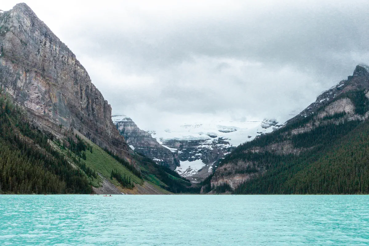 Turquoise water at Lake Louise with mountains and a glacier in the background in Banff National Park, Canada. Forested slopes and rocky cliffs surround the lake under a cloudy sky.