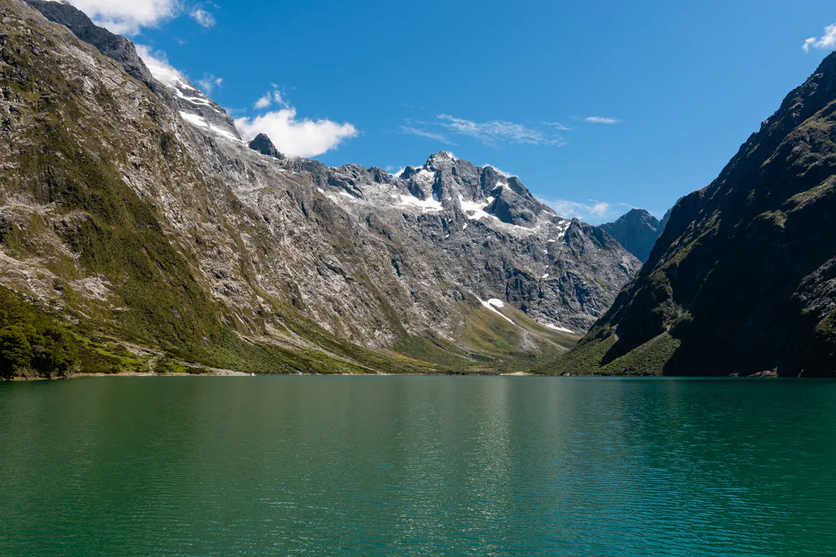 Lake Marian, a beautiful alpine lake in Fiordland National Park, is surrounded by steep rocky mountains with patches of snow and lush green vegetation. The calm green lake sits in a remote alpine valley.