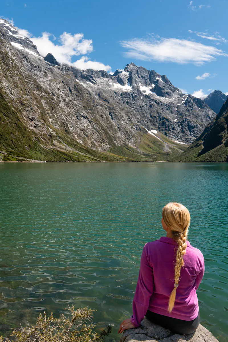 A hiker sitting on a rock at the edge of Lake Marian in Fiordland National Park, New Zealand, looking out over the calm green alpine lake surrounded by steep rocky mountains with patches of snow under a bright blue sky.