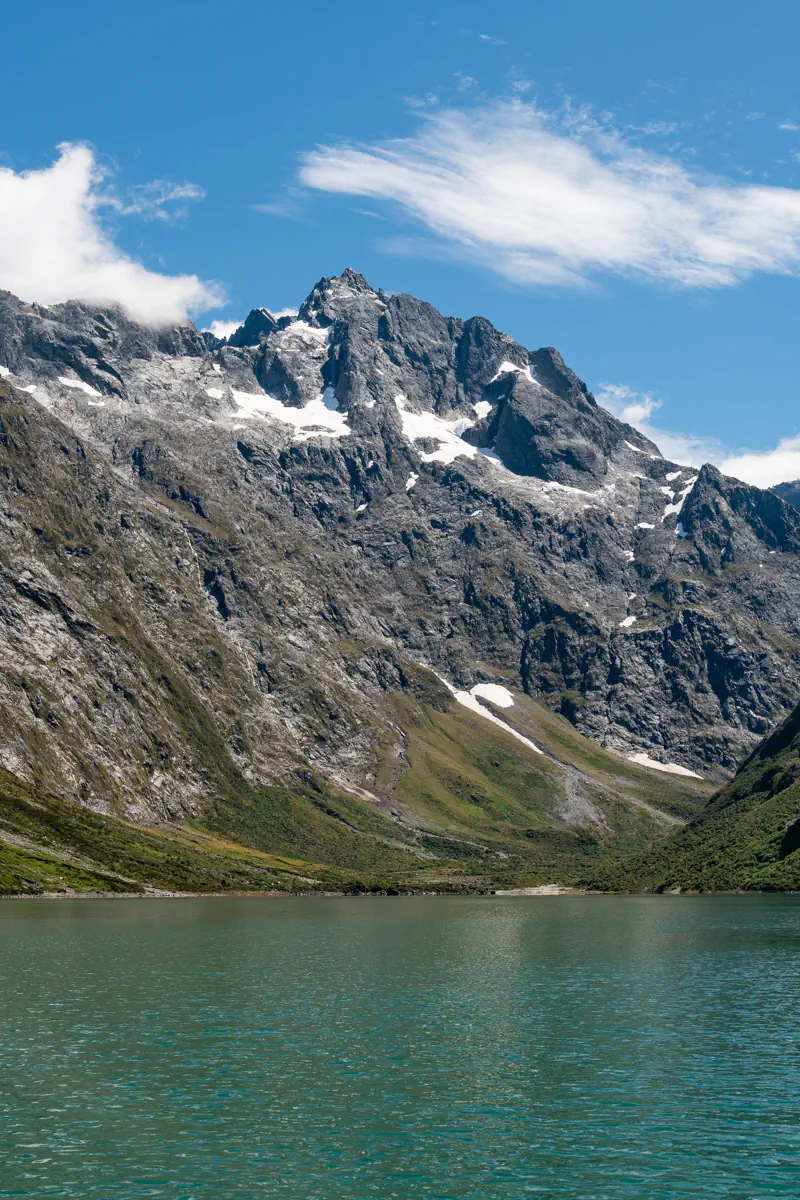 Lake Marian in Fiordland National Park surrounded by steep rocky mountains with patches of snow. The calm green alpine lake stretches across the valley below the rugged peaks.