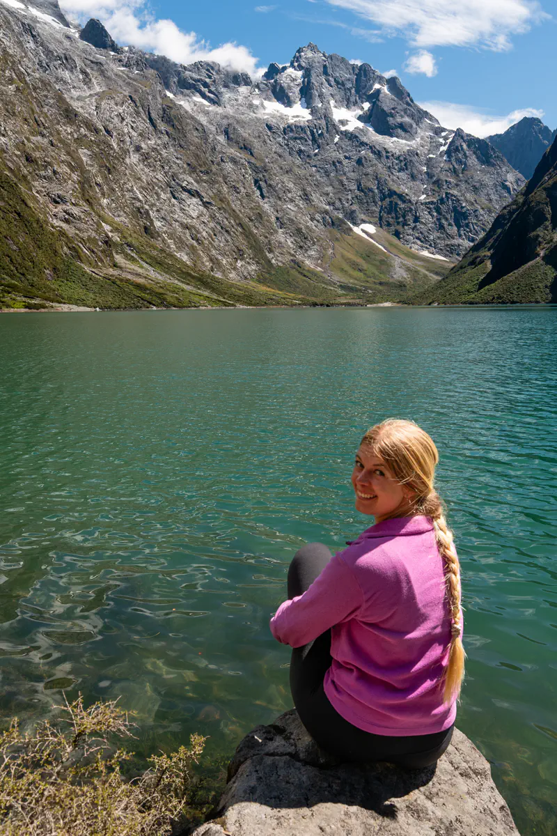 Person sitting on a rock at the edge of Lake Marian with steep rocky mountains and patches of snow rising behind the lake.