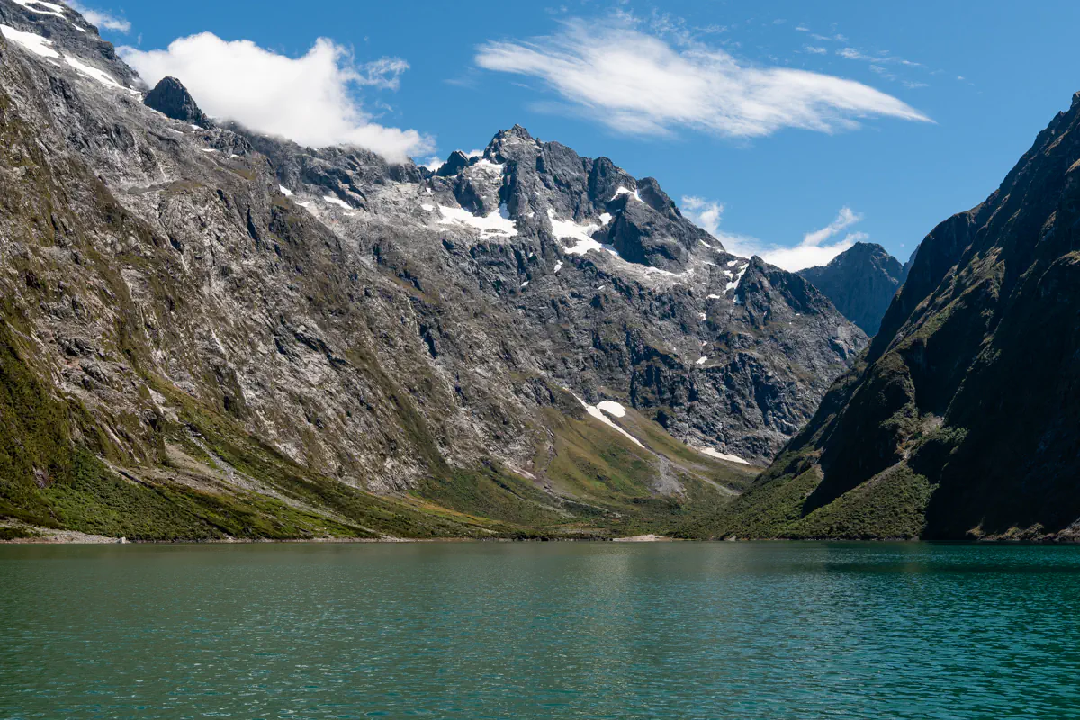 Lake Marian in Fiordland National Park, a clear emerald alpine lake surrounded by steep rocky mountains, rugged slopes, and patches of snow.