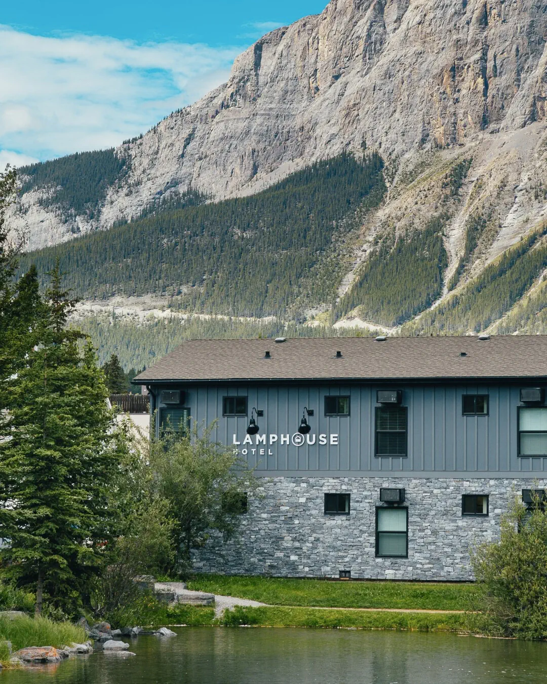 Lamphouse Hotel building in Canmore, Alberta with the Rocky Mountains rising behind it. The hotel sits beside a small pond with trees and forested slopes leading up to the rocky mountain cliffs.