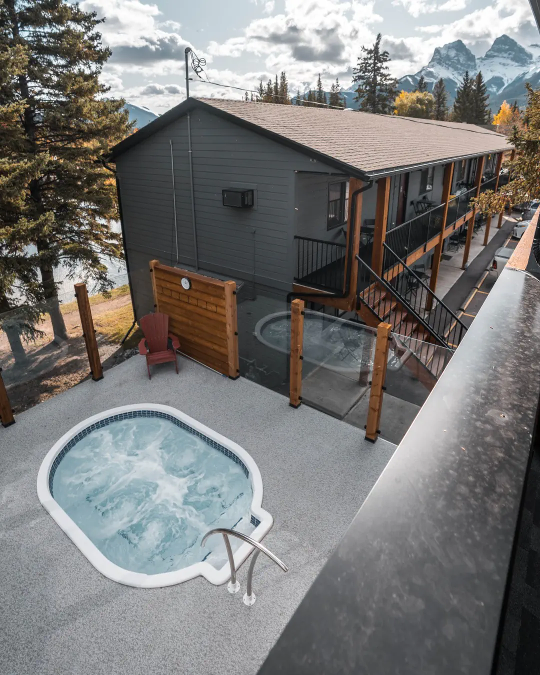 Outdoor hot tub at a hotel in Canmore, Alberta with views of the surrounding Rocky Mountains. The two-story lodge building is surrounded by trees with mountain peaks visible in the background.