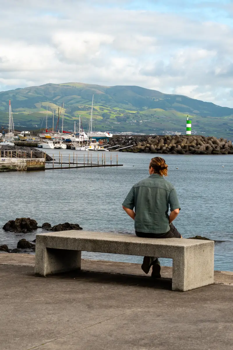 A man sits on a concrete bench overlooking a small harbour with sailboats and a rocky breakwater. Rolling green hills rise in the background under a cloudy sky.