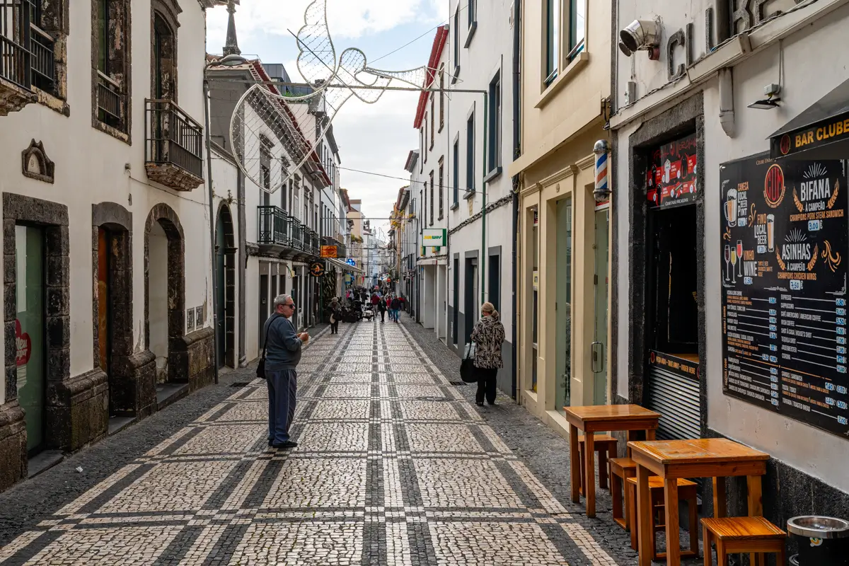 A narrow cobblestone street lined with shops and restaurants in Ponta Delgada, with people walking along the patterned pavement. Outdoor seating and signage line the buildings on either side.