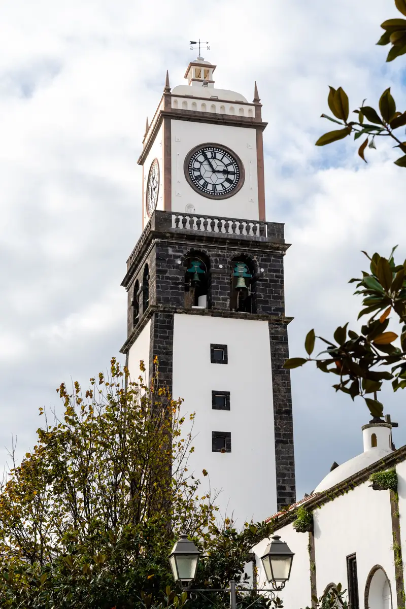 A tall clock tower with white walls and dark stone trim rises above nearby buildings and trees. The clock face and bell openings are visible against a partly cloudy sky.