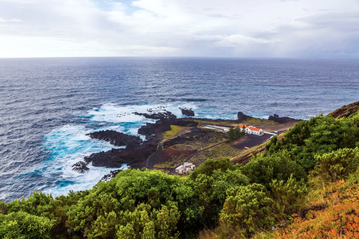 A coastal viewpoint in São Miguel, Azores overlooking dark volcanic rocks and waves crashing along the shoreline. A small white building with a red roof sits near the water, surrounded by green vegetation on the hillside.