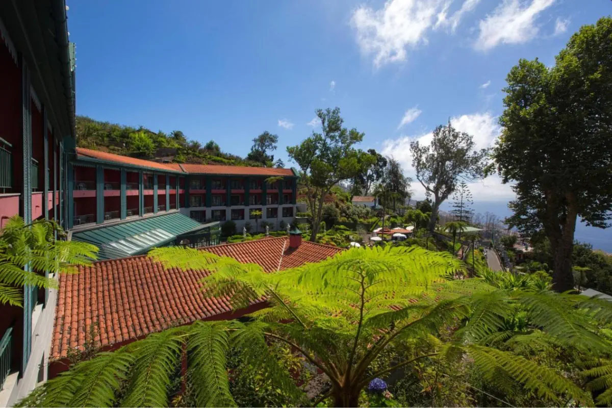 A hillside hotel with red-tiled roofs and green balconies is surrounded by lush tropical plants and tall trees under a bright blue sky. The ocean is visible in the distance beyond the landscaped gardens and terraces.