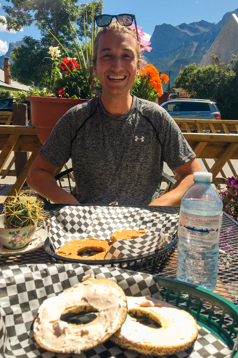 A smiling man sitting at an outdoor table with bagels and drinks, with flowers and mountains in the background.