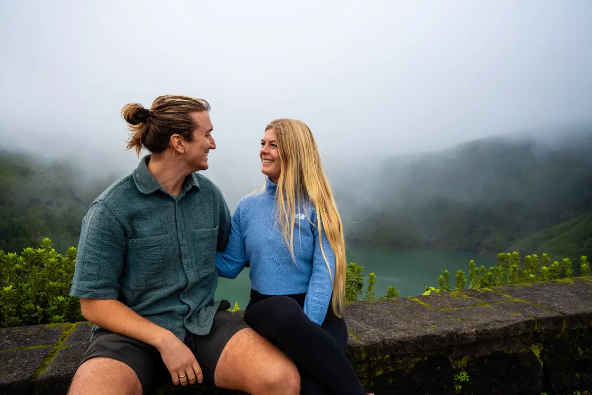 A couple sitting on a stone wall overlooking a misty crater lake surrounded by green hills in São Miguel, Azores.