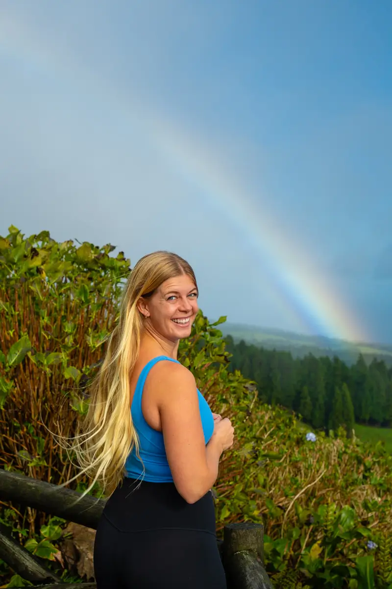 A woman standing by a wooden railing with a rainbow visible over a lush green valley in the Azores.