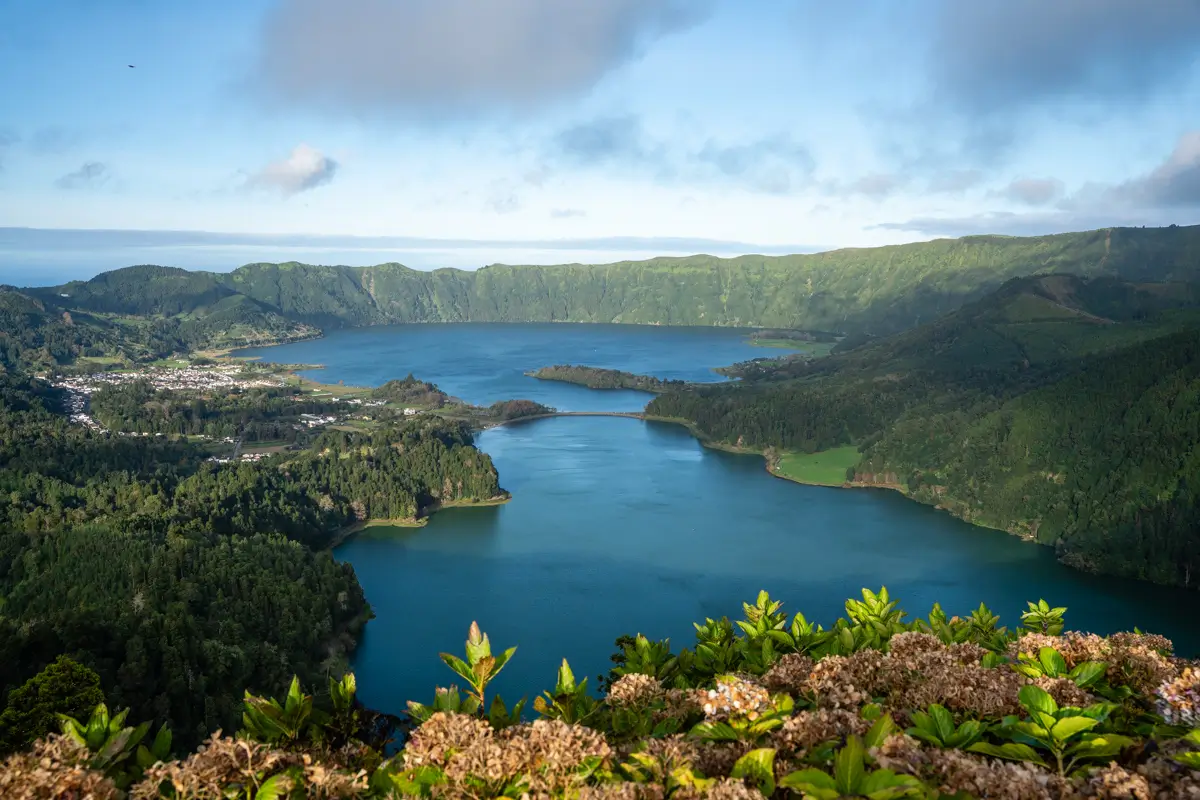 View over Sete Cidades in São Miguel, Azores, showing twin crater lakes separated by a narrow bridge and surrounded by steep green hills. The deep blue water contrasts with lush forest and a small village along the shoreline.