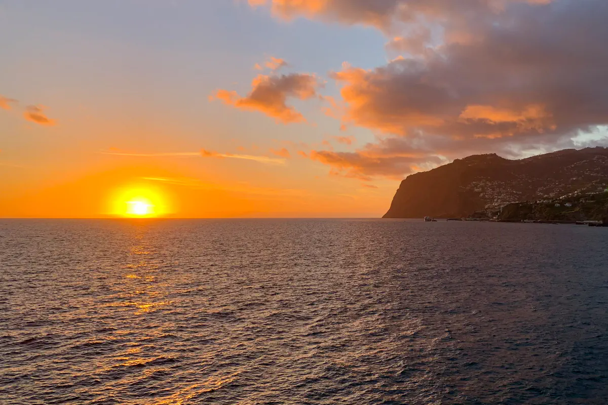 The sun sets over the ocean, casting golden light across the water as a rocky cliff and hillside town appear in the distance. Warm orange and pink clouds fill the sky above the coastline.